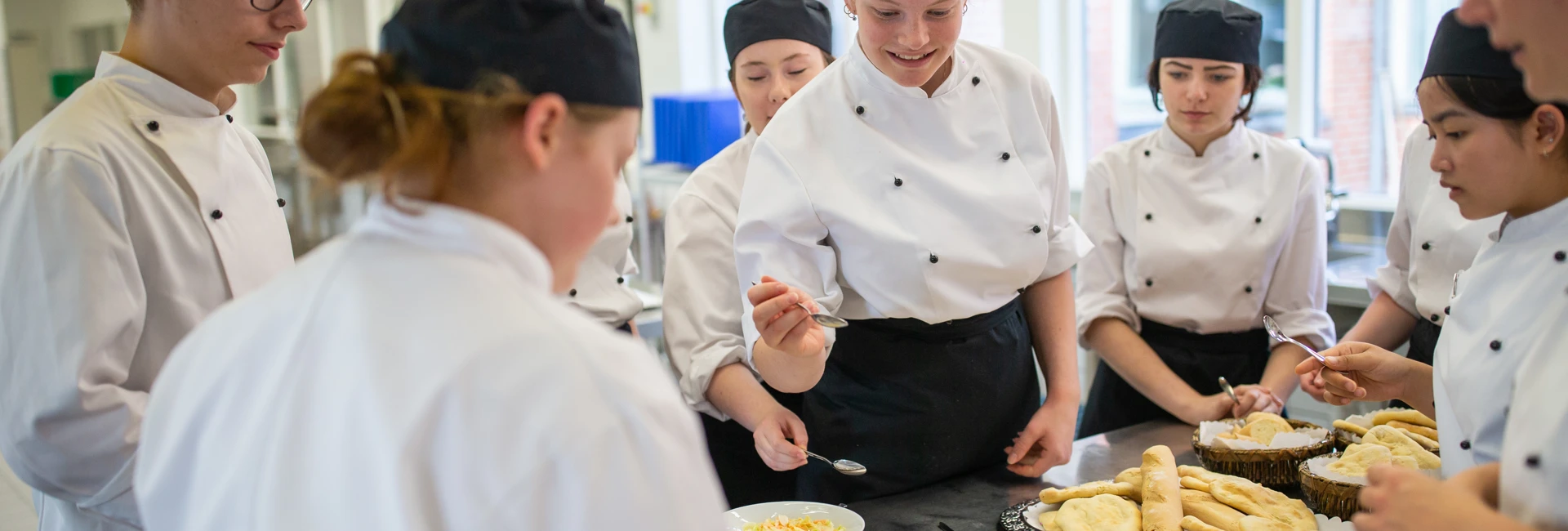 Elever i kokkeuniform samles om et bord og laver mad